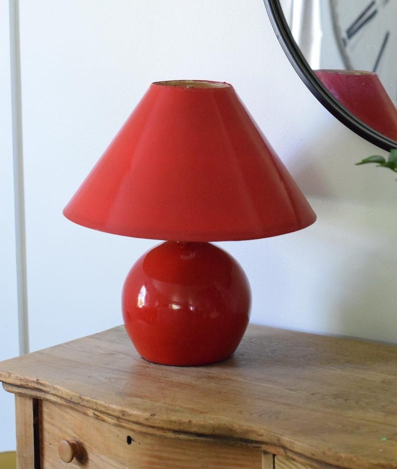 May include: A red table lamp with a round base and a conical shade. The lamp is glossy and sits on a wooden surface. A round mirror is partially visible in the background, reflecting the lamp and a clock face. The lamp is a decorative lighting fixture.