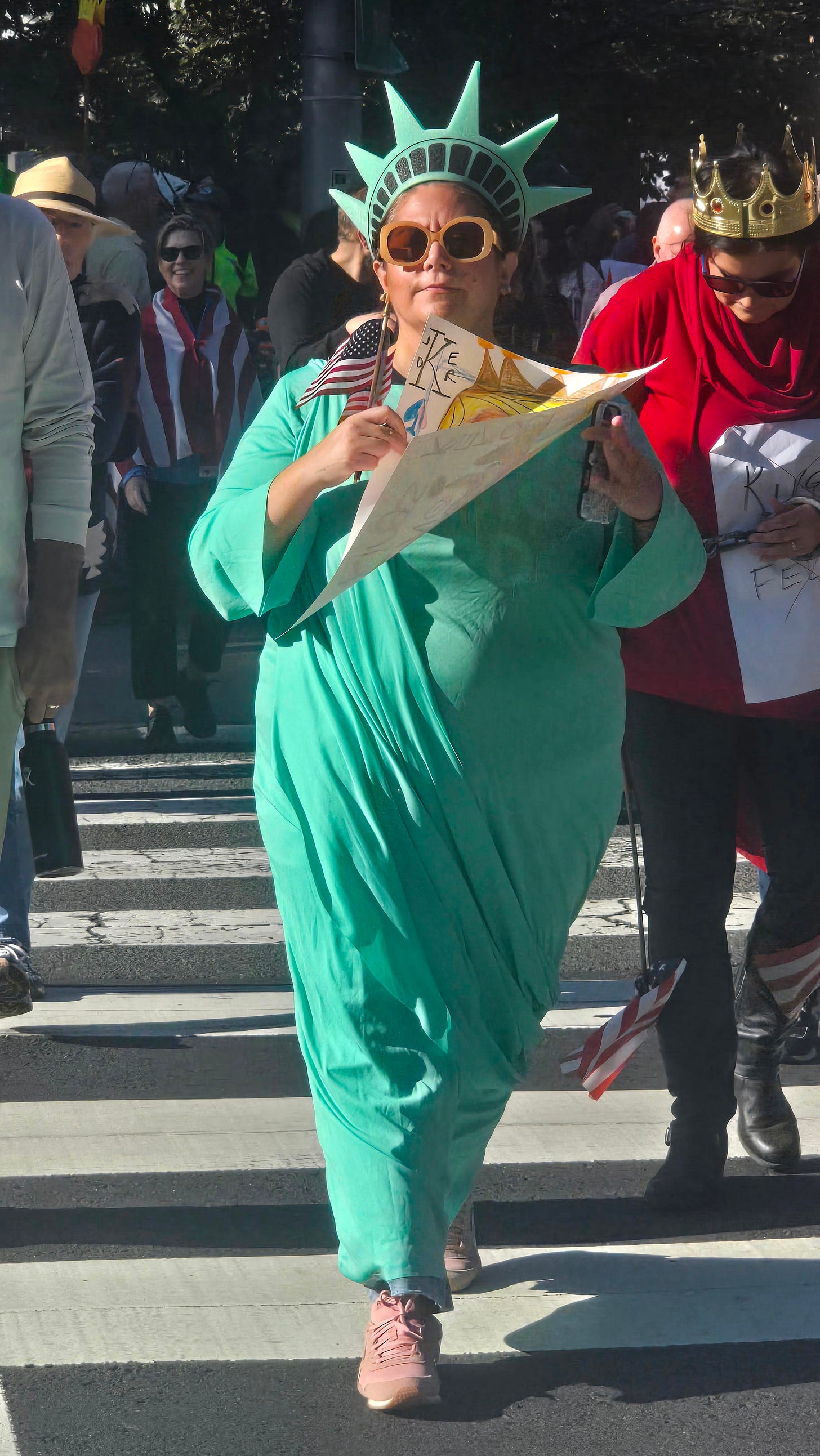 a woman walking down the street dressed as the statue of Liberty holding an American flag and a sign, wearing sunglasses and sneakers a woman walking down the street dressed as the statue of Liberty holding an American flag and a sign, wearing sunglasses and sneakers