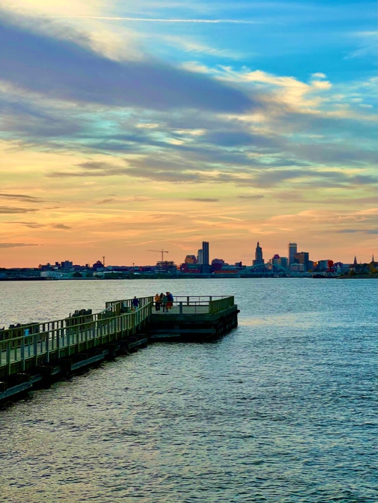 Sunset over Kettle Point Pier in Providence, Rhode Island, showing the abandoned railway bridge extending into Narragansett Bay, with silhouetted industrial cranes and golden clouds in the background — a haunting, tranquil coastal view blending natural beauty and forgotten infrastructure.