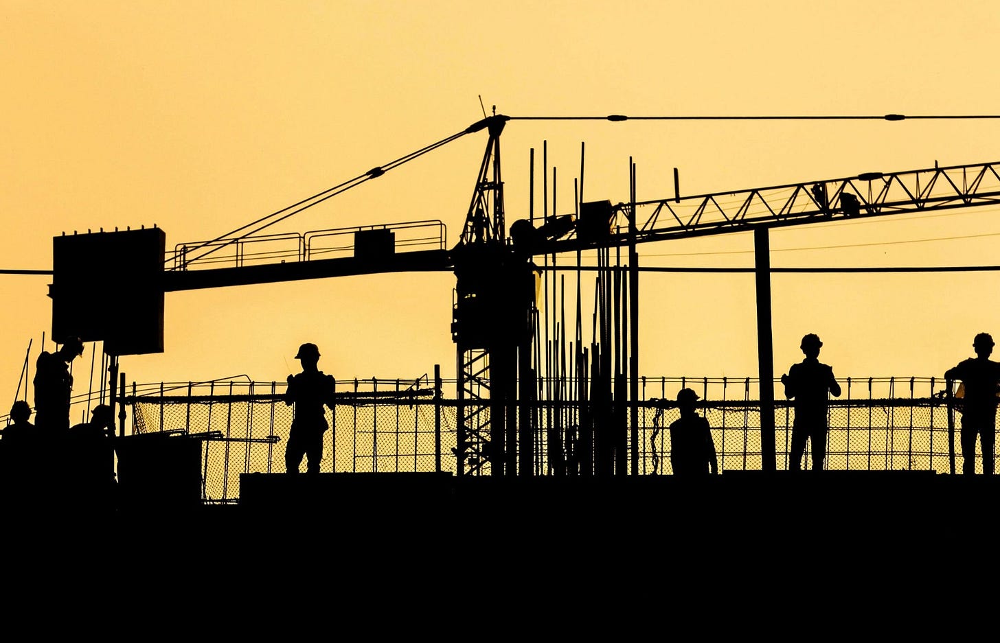 Construction site silhouette with workers, cranes, and steel framework against a yellow sky.