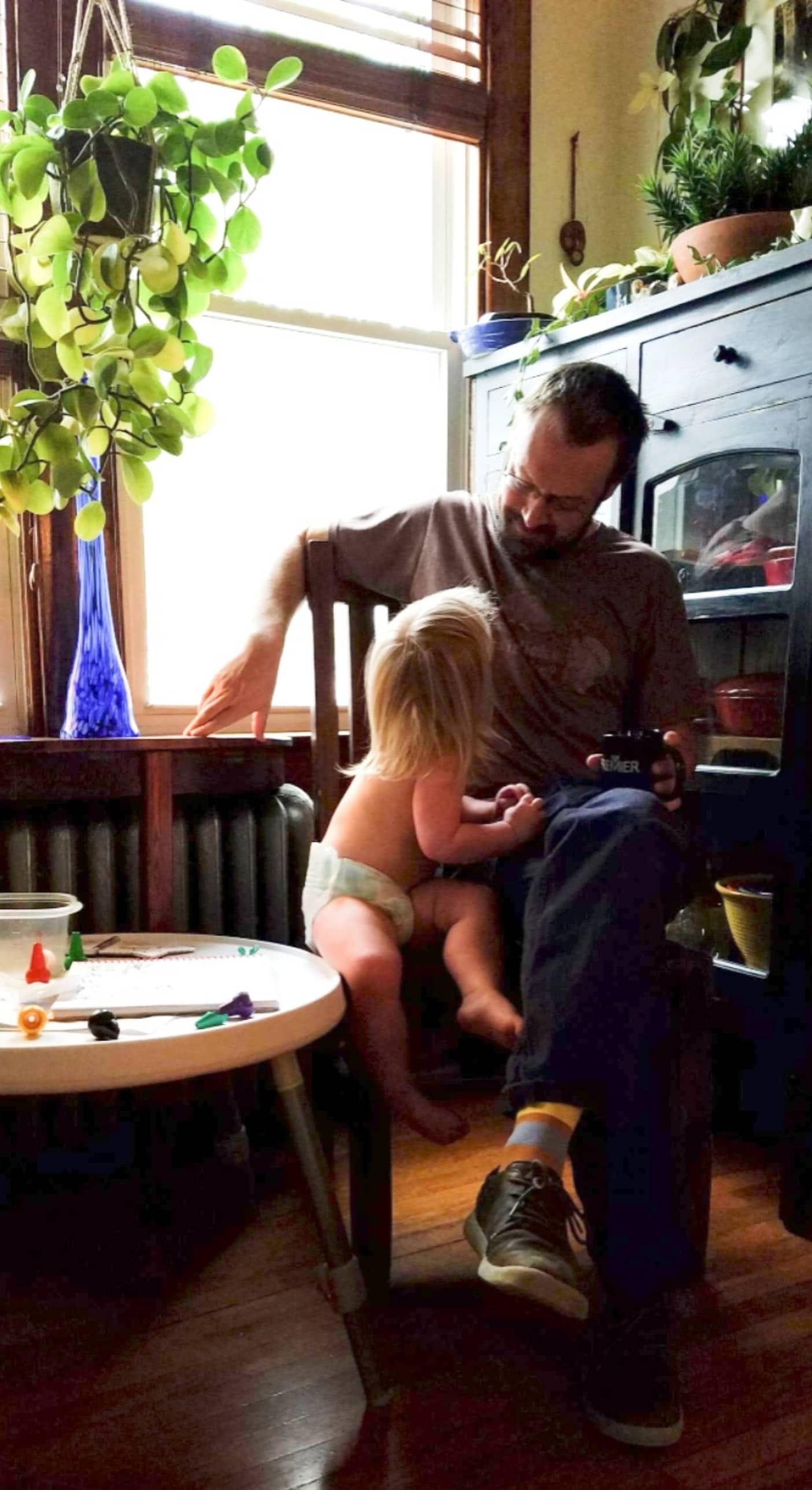 Photo of a man sitting in a wooden chair with a small child looking at each other. The scene is indoors under a sunlit window with plants all around.