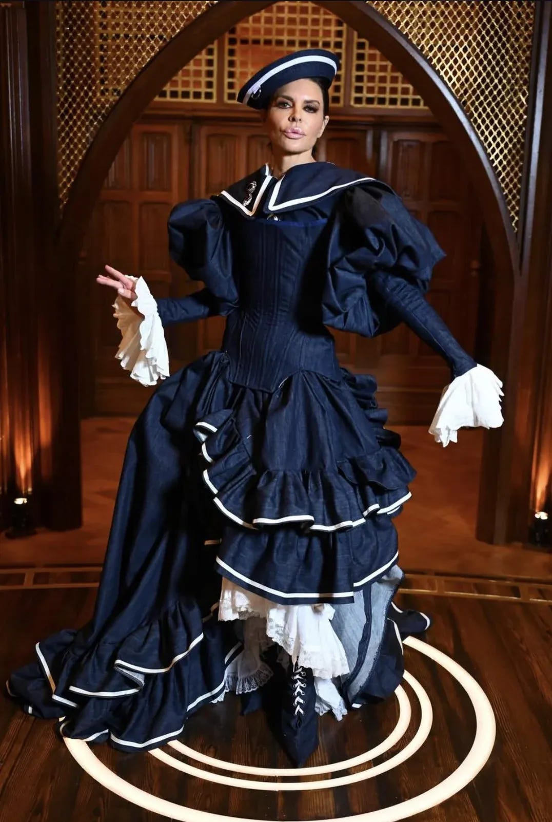 Lisa in a navy Victorian-inspired gown with white trim, corseted waist, tiered ruffled bustle skirt, exaggerated puff sleeves, and a matching sailor-style hat.