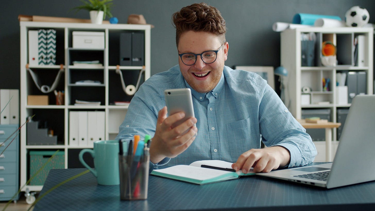 Cheerful man in glasses using smartphone at a desk with laptop. Cheerful man in glasses using smartphone at a desk with laptop.