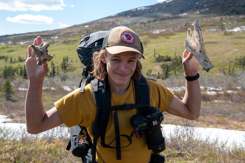 A young man in a yellow shirt and ball cap, a camera slung across his chest, grins as he holds up a weathered bone in each hand, green tundra and snowy slope behind.