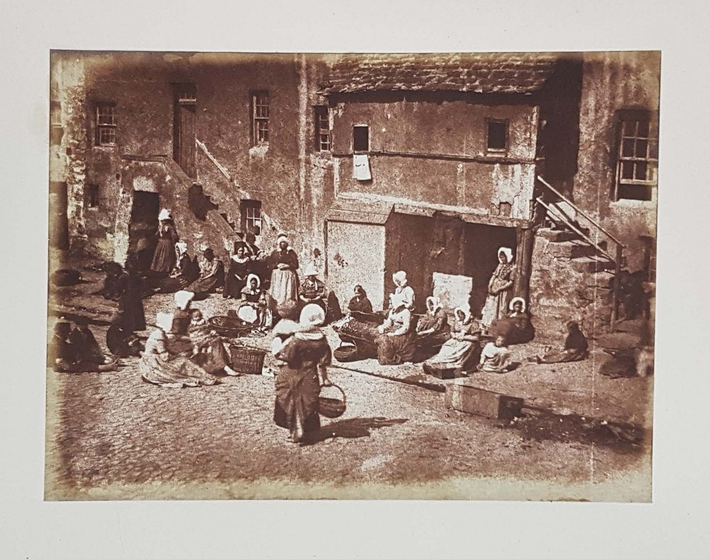 Photographic print showing women seated in front of houses preparing fishing lines. A woman holding a baby approaches them.
