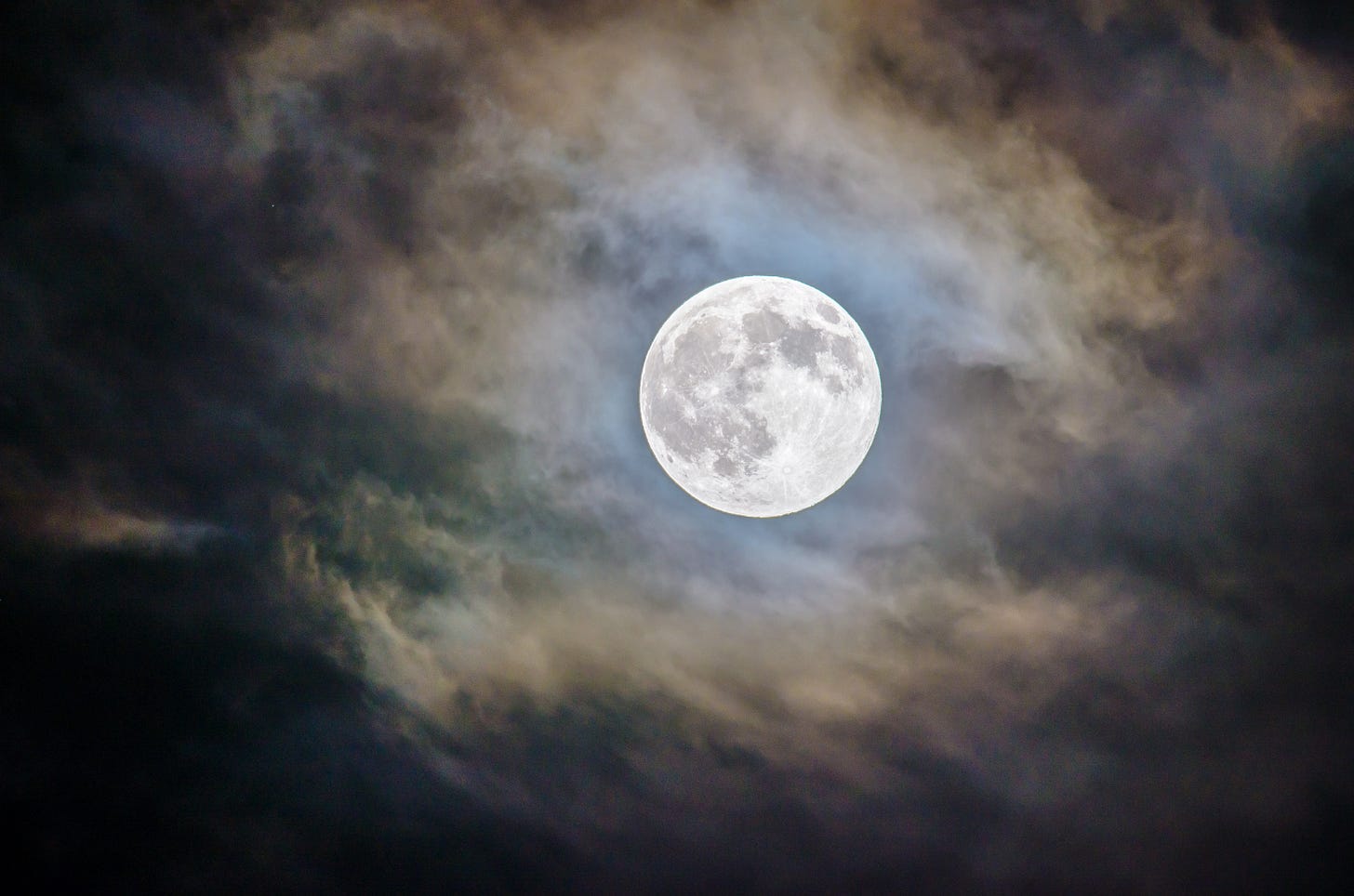 A photo of the full moon in the night sky, seen through a thin layer of wispy moonlit clouds A photo of the full moon in the night sky, seen through a thin layer of wispy moonlit clouds