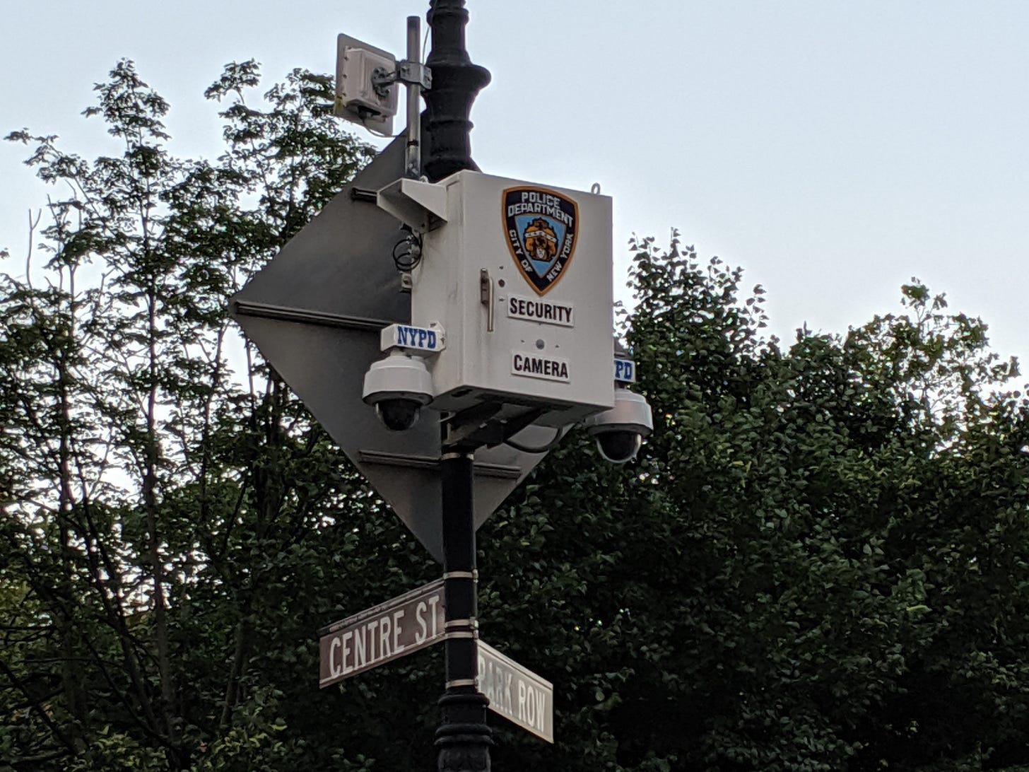 A photo of a white box with two protruding surveillance cameras attached to the opposite sides of the box. The box is attached to a lamp post and is marked with "NYPD," "Security Camera," and the NYPD's shield. Below the surveillance box and cameras are street signs for Centre St. and Park Row.