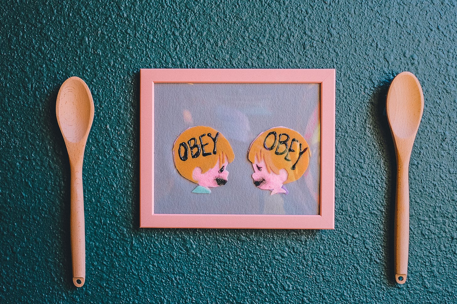 inside a pink frame two felt child who look a bit like precious moments characters have zombie mouths and hte words obey in their heads. the picture is surrounded by two wooden spoons