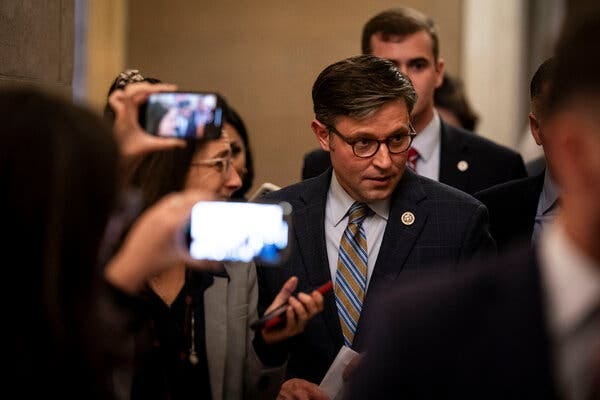 Speaker Mike Johnson wearing a dark suit, blue shirt and striped tie. Reporters hold their phones and surround him.