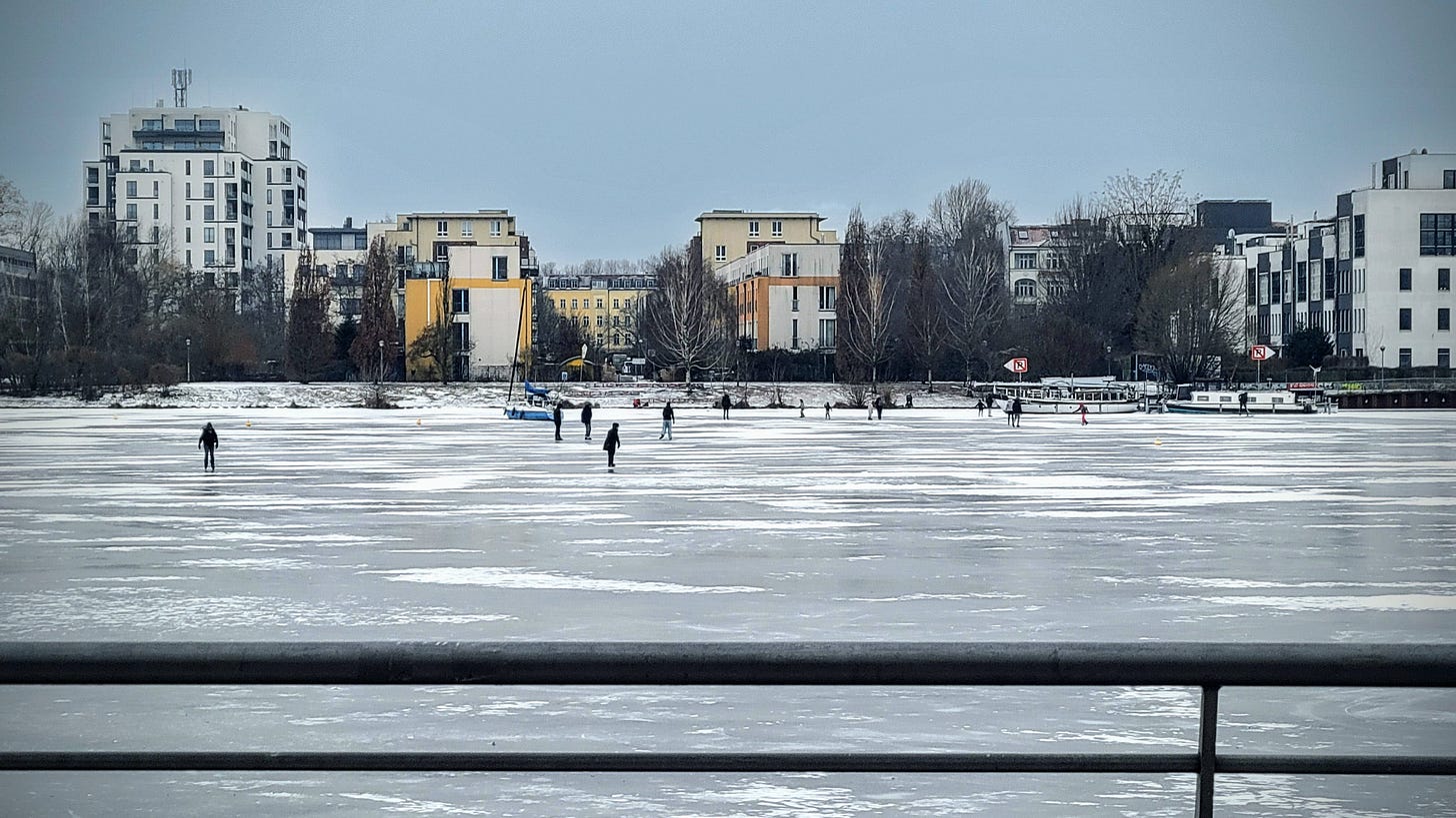 Foreground: Metal railing runs along the bank of a large frozen lake. In the distance, idle boats wait for a thaw and individual people are skating on the ice. Skyscrapers rise in the background on the opposite shore.