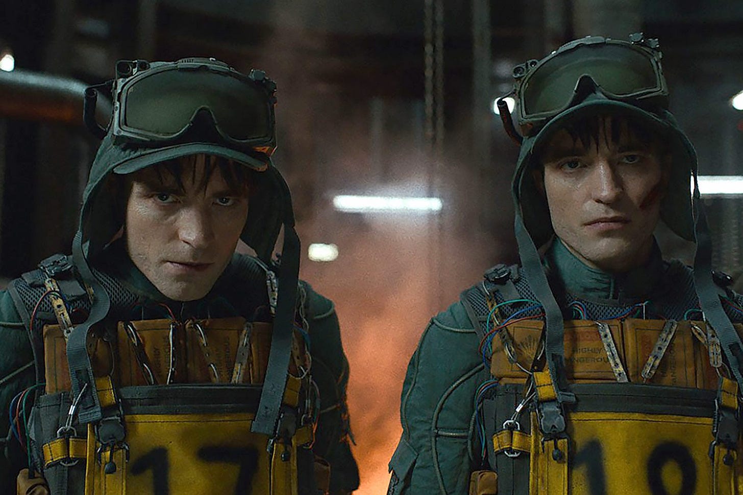 Two young men in protective suits and goggles in a factory environment with flames behind them.