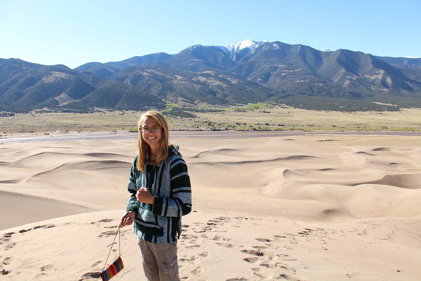 a young lady holding a small purse, standing on sand dunes with a mountain range behind her