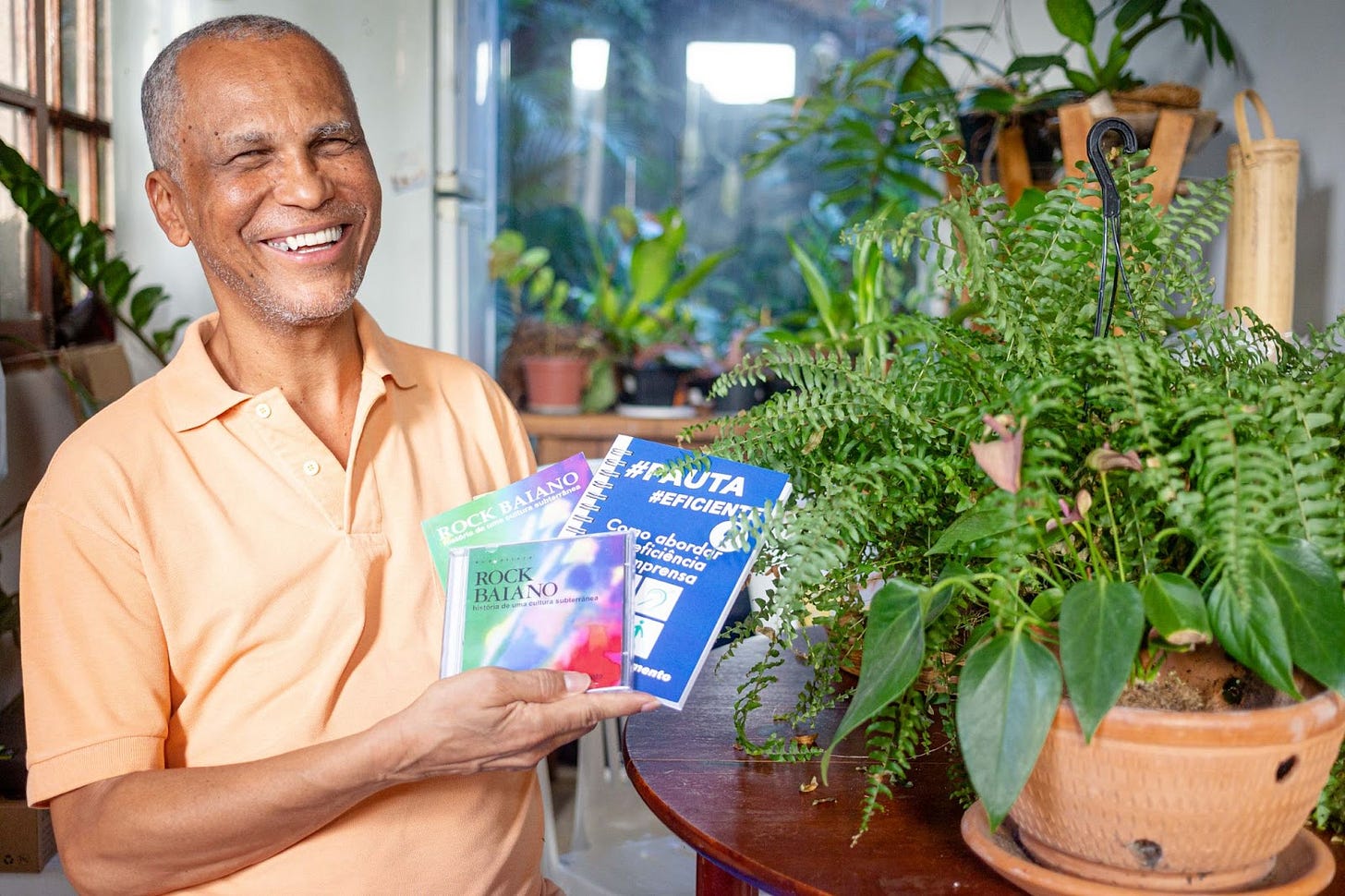 Foto com cores vibrantes com Ednilson sentado, em plano médio, ao lado esquerdo da imagem, sorrindo. Ele é negro de pele clara, cabelos curtos e grisalhos, está usando uma blusa de cor salmão e segura os CD e livro Rock Baiano junto com o livro Pauta Eficiente. Carrega ambos com uma das mãos. Ao seu lado tem uma série de plantas, de tom verde escuro em vasos de barro. Elas estão sobre uma mesa de madeira