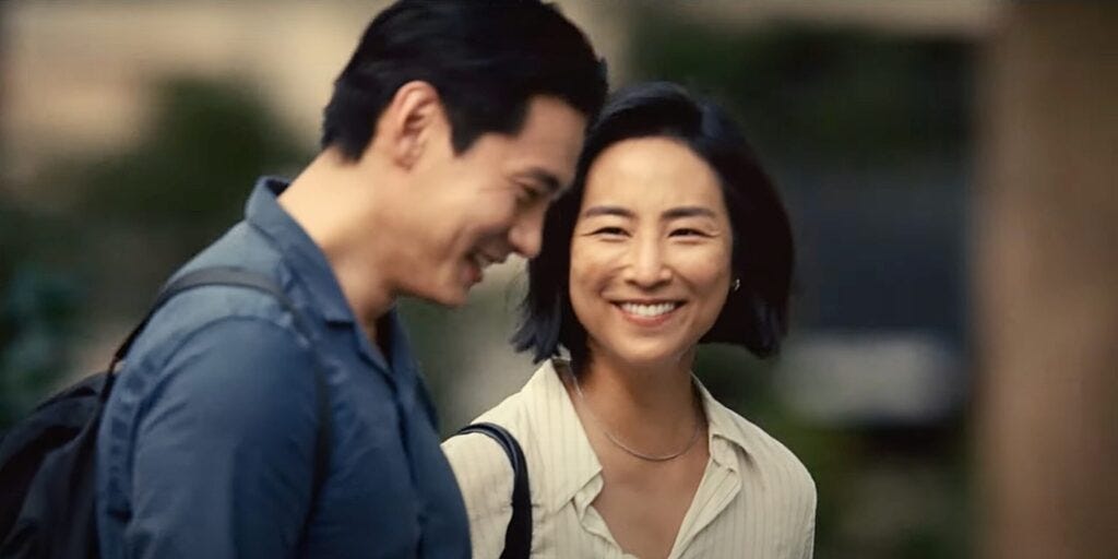 A Korean-Canadian woman with a black haired bob in a cream-striped collared shirt, is in focus, smiling at the black-haired Korean man ahead in front of her. He is looking down, and from the side profile, he is smiling as well. He is wearing a navy blue button-up dress shirt, and has a black backpack on.