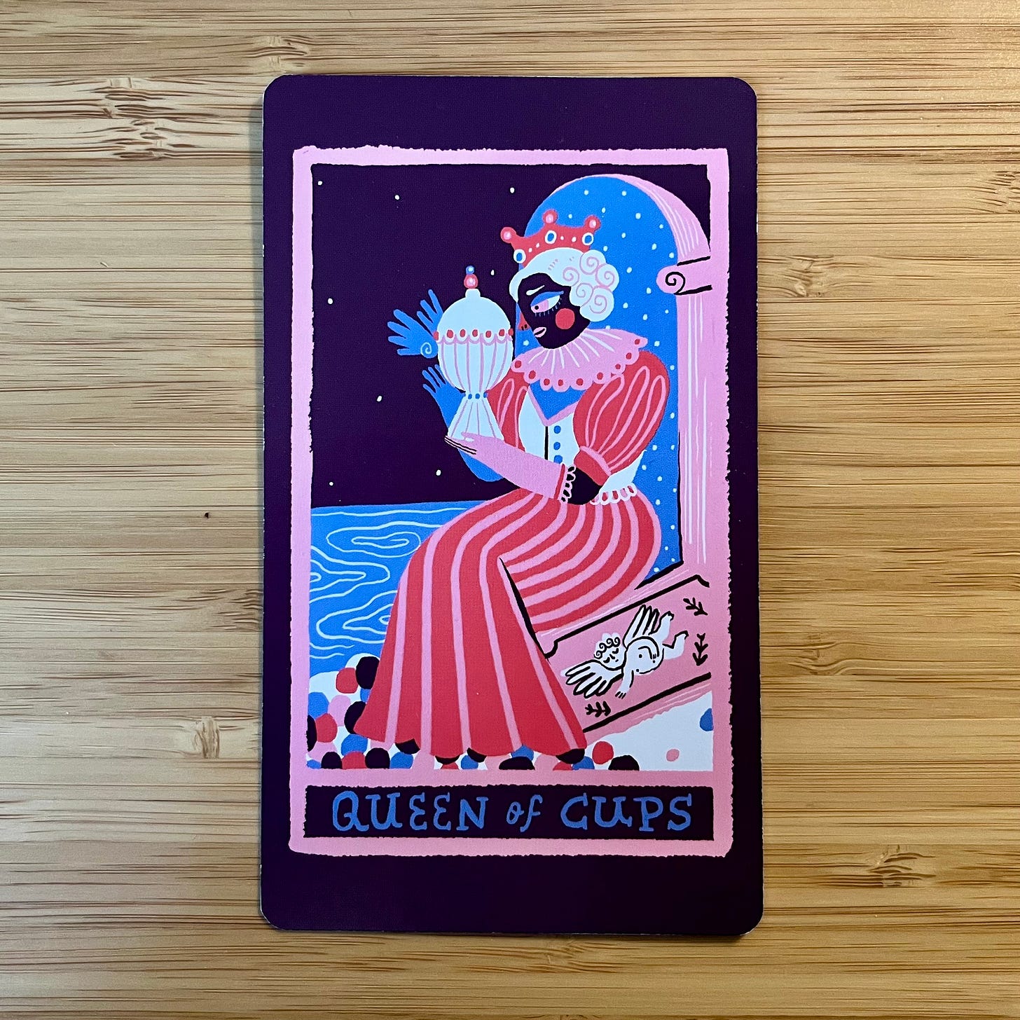 An image of a tarot card with a dark, eggplant purple background. The main part of the tarot image is framed within a bubblegum pink rectangular border that looks like it was drawn with crayon. At the bottom of the rectangle the text "Queen of Cups" is written in blue, capital letters. Within the border a regal figure sits on a throne in profile, holding and looking at a white vessel with red, pink, and blue decorative details that also appears to have water splashing out of it. The Queen's hair is white, she's wearing a pink and red striped dress, and she has on one pink glove and one blue glove. At her feet are blue, red, pink, and purple stone-like shapes. In the background, a blue body of water with gentle eddies. The back of the throne is an optical illusion: an archway through which you can see a blue sky speckled with tiny white stars. A small Cupid figure is painted on the side of the throne in white, surrounded by three black arrows. An image of a tarot card with a dark, eggplant purple background. The main part of the tarot image is framed within a bubblegum pink rectangular border that looks like it was drawn with crayon. At the bottom of the rectangle the text "Queen of Cups" is written in blue, capital letters. Within the border a regal figure sits on a throne in profile, holding and looking at a white vessel with red, pink, and blue decorative details that also appears to have water splashing out of it. The Queen's hair is white, she's wearing a pink and red striped dress, and she has on one pink glove and one blue glove. At her feet are blue, red, pink, and purple stone-like shapes. In the background, a blue body of water with gentle eddies. The back of the throne is an optical illusion: an archway through which you can see a blue sky speckled with tiny white stars. A small Cupid figure is painted on the side of the throne in white, surrounded by three black arrows.