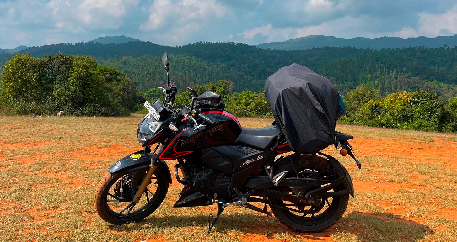A black TVS Apache motorcycle is parked on a grassy field in front of a range of lush, green mountains. The motorcycle has red accents and is carrying a covered package on the back. The sky is partly cloudy. A black TVS Apache motorcycle is parked on a grassy field in front of a range of lush, green mountains. The motorcycle has red accents and is carrying a covered package on the back. The sky is partly cloudy.