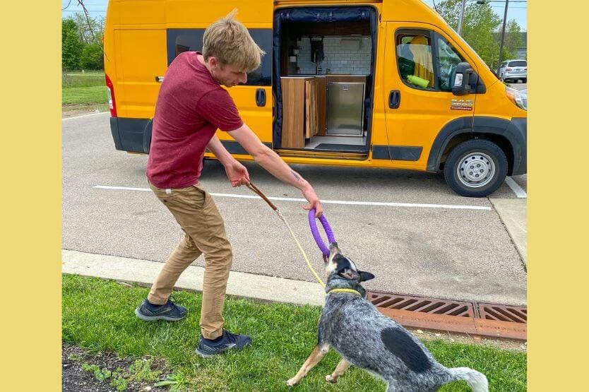 Scout the blue heeler plays tug with her owner Sean, a young man, with a purple puller toy in front of our parked converted camper van