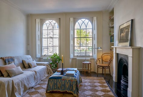 A wooden bay window with blue seat, a soft pink dining room with antique table, a small living room with a sofa and ottoman