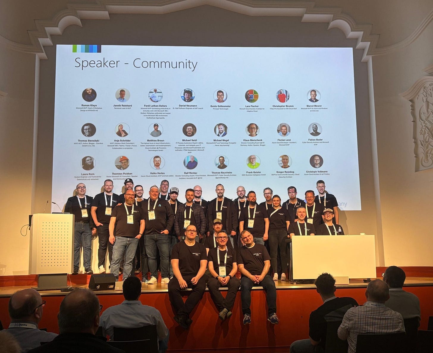 Hier ist ein geeigneter **Alt-Text** für das Bild:  **Alt-Text:**   Gruppenfoto der Speaker auf der Bühne der Experts Live Germany 2025 in Leipzig. Im Hintergrund eine große Leinwand mit den Namen und Profilbildern der Vortragenden. Die Bühne ist in einem historischen Saal mit weißem Stuckdekor. Die Speaker tragen überwiegend schwarze T-Shirts mit Event-Badges. Einige sitzen vorn am Bühnenrand, andere stehen dahinter. Publikum ist im Vordergrund zu sehen.