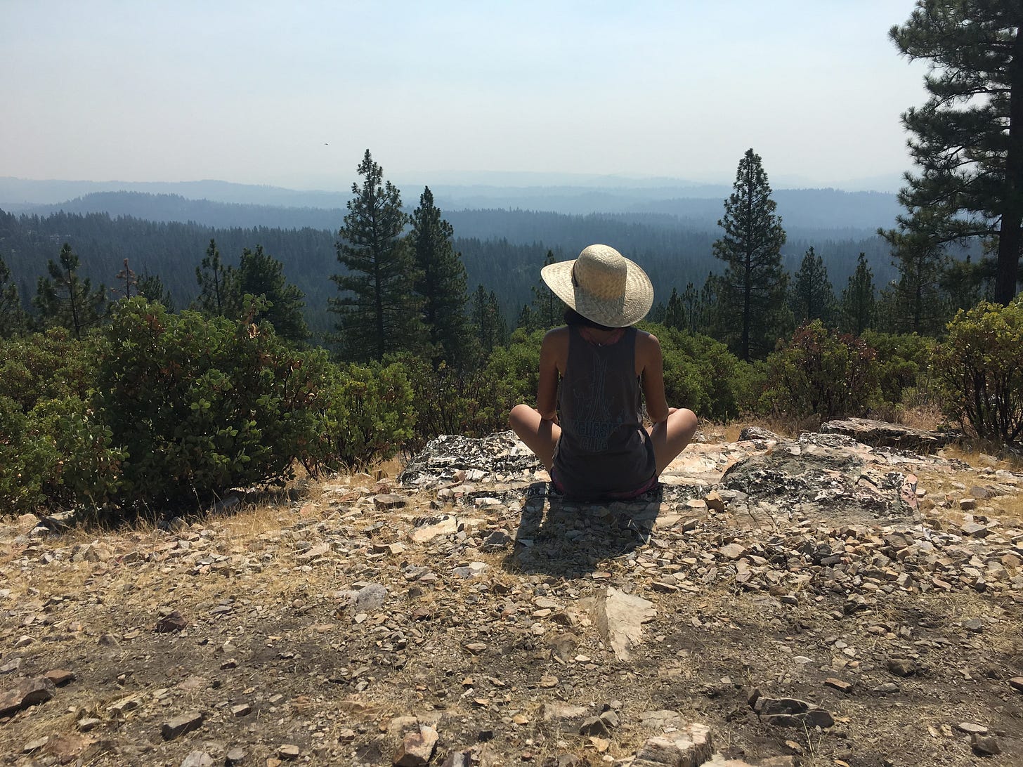 A brown-skinned woman, shown from behind. She wears a wide-brimmed hat, gray tank top, and black shorts. She sits on a rocky dirt area, overlooking an expansive viewpoint full of evergreens.