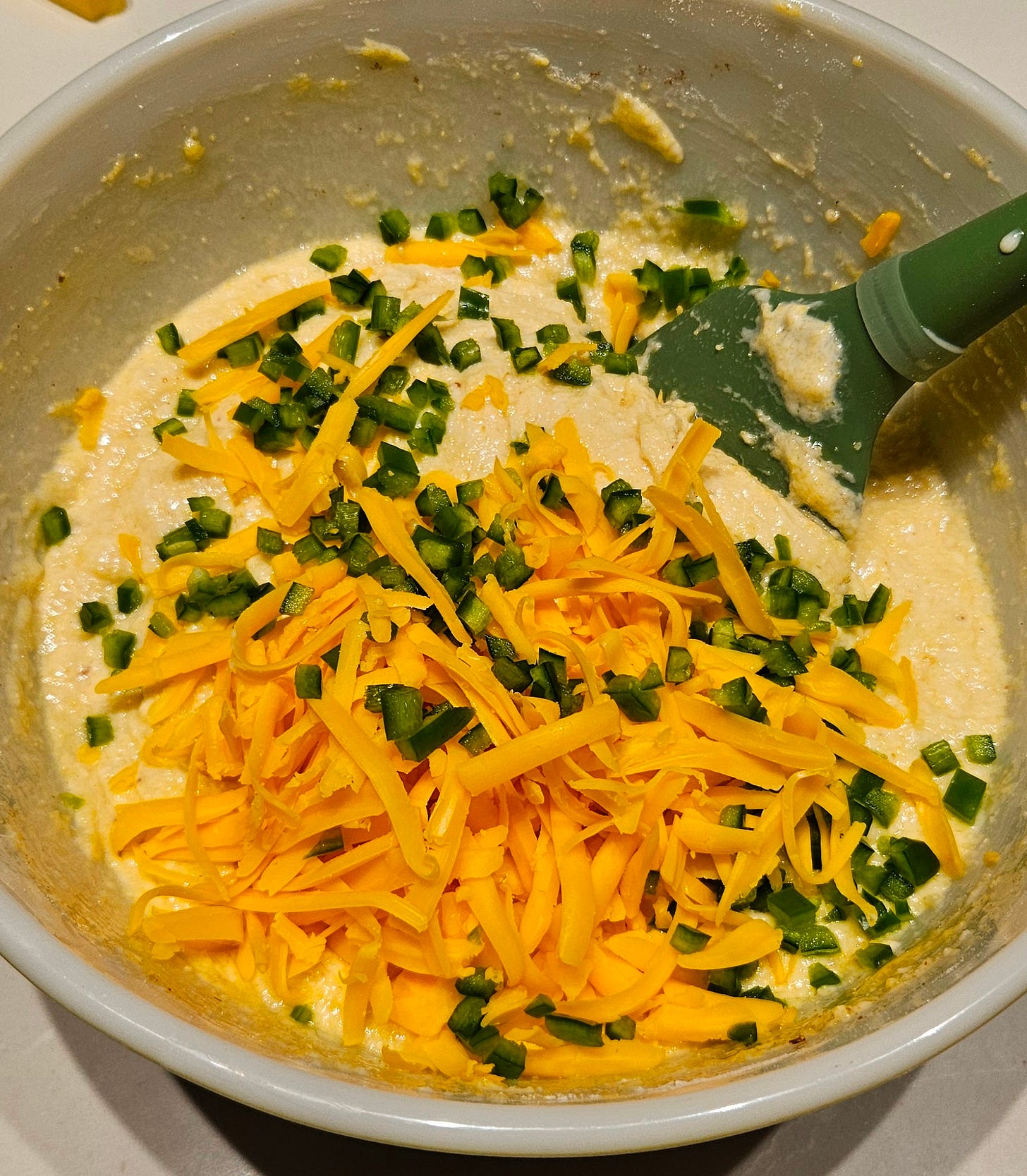 Shredded cheese and diced jalapenos being added to the corn bread batter with a green spatula