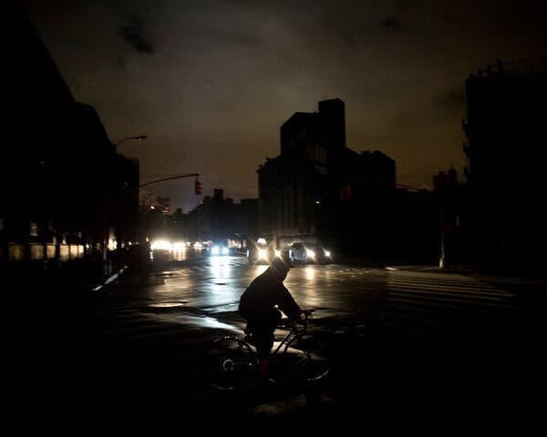 A biker crosses a street that is almost pitch black, except for the headlights of oncoming cars, at night. A biker crosses a street that is almost pitch black, except for the headlights of oncoming cars, at night.