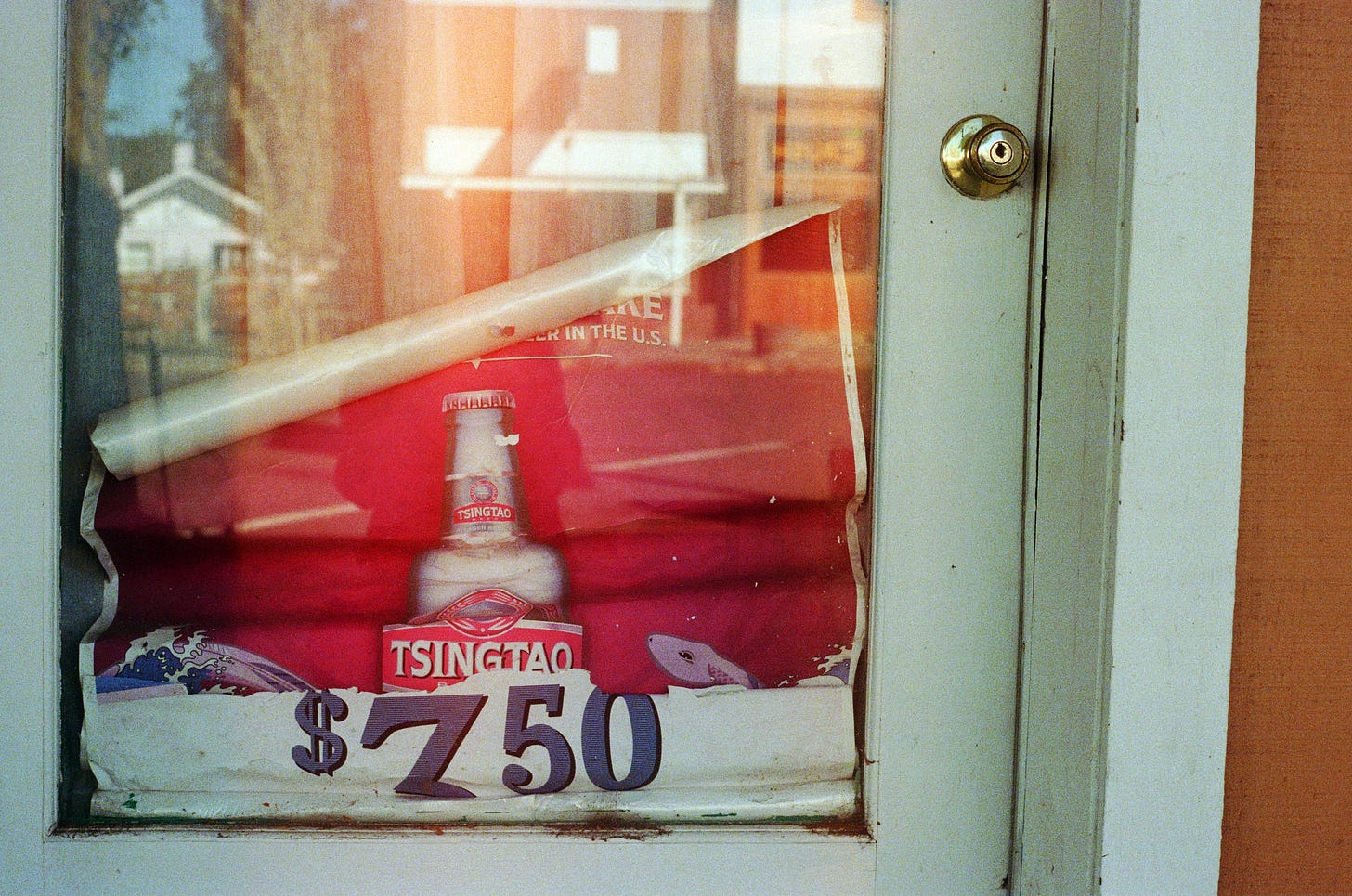 A close-up film photo of a door, where an old advertisement for Tsing Tao, discolored from sun exposure, is crumbling apart and falling down the door's glass center.