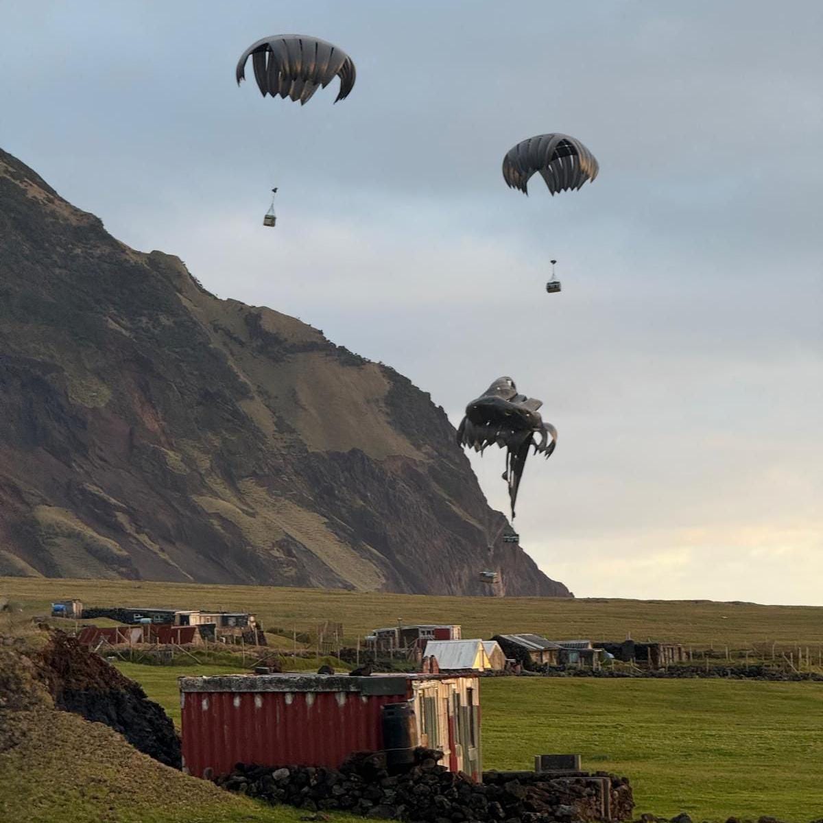 Parachutes delivering boxes of military supplies seen against a rugged backdrop, where several small buildings can be seen Parachutes delivering boxes of military supplies seen against a rugged backdrop, where several small buildings can be seen