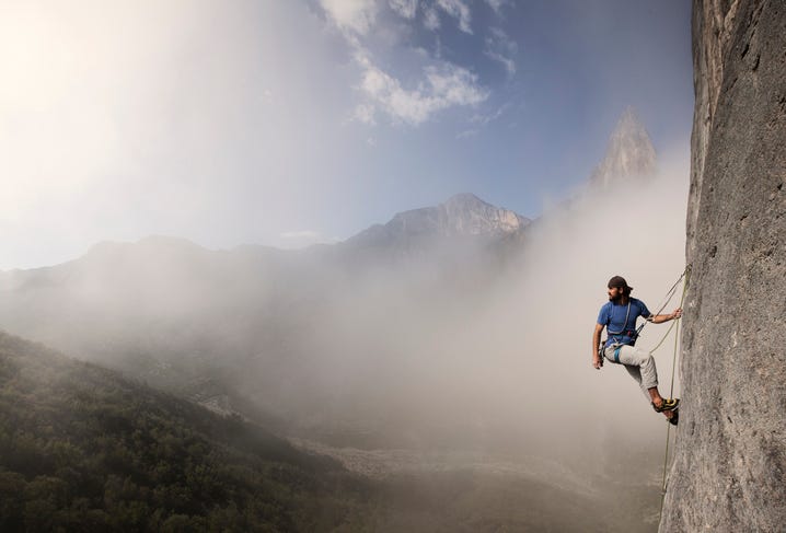 a man in a blue shirt and white pants is paused in the middle of climbing a mountain. He is looking outward to the scene below him--trees, mountains, a blue sky.