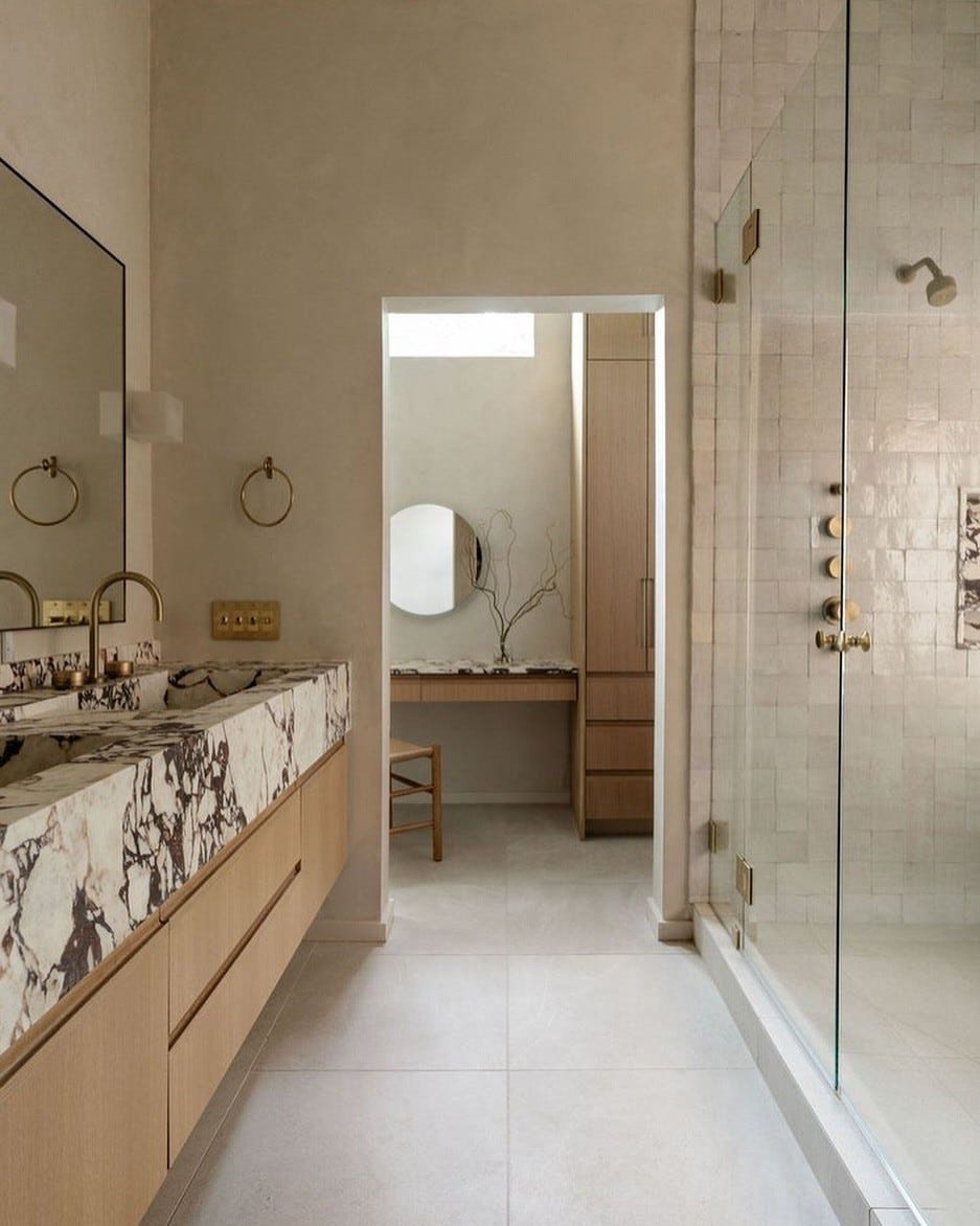 calming bathroom with marble counter tops and cream tiles directing you to a dressing area creating a calm space for self care.