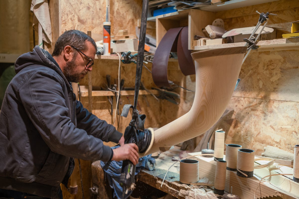Sandro Faïta shaping the bell of an alphorn with a power tool in his woodshop, surrounded by materials and equipment.