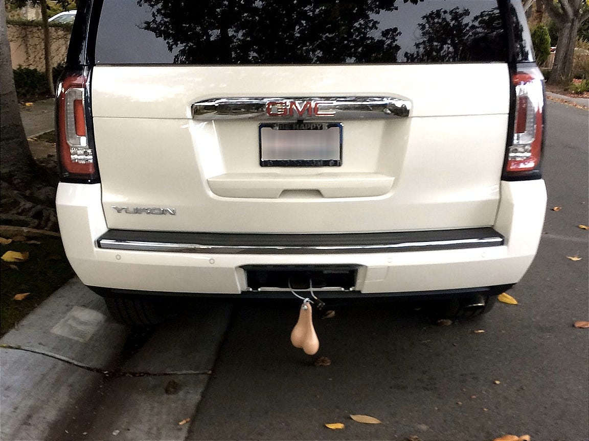 Rear view of a white GMC Yukon parked on a residential street, with a pair of flesh-colored novelty “truck nuts” hanging from the trailer hitch.