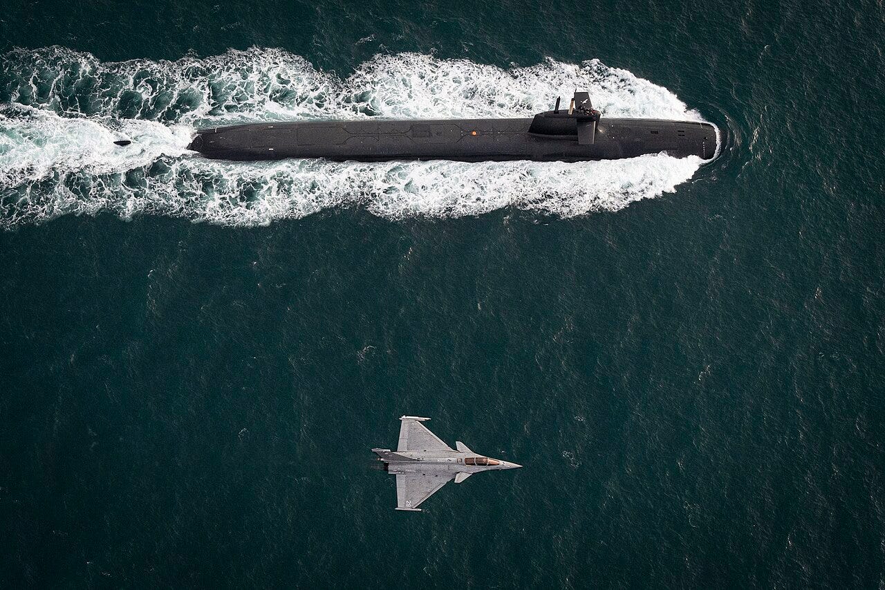 Overhead view of a French ballistic missile submarine on the surface leaving a white wake, with a Rafale fighter jet flying low over the dark water below it. Overhead view of a French ballistic missile submarine on the surface leaving a white wake, with a Rafale fighter jet flying low over the dark water below it.