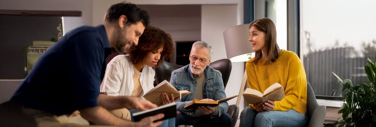A imagem mostra quatro pessoas sentadas em um ambiente interno, participando de um momento de leitura ou estudo em grupo. Elas estão em cadeiras confortáveis, formando um círculo informal, e cada uma segura um livro aberto.  À esquerda, um homem de cabelos escuros e barba veste uma camisa azul escura, inclinando-se levemente para frente enquanto lê. Ao seu lado, uma mulher negra de cabelo cacheado usa uma camisa branca sobre uma camiseta bege e também está concentrada na leitura. Ao centro, um homem idoso, de cabelo grisalho e óculos, veste uma jaqueta jeans e observa o livro em suas mãos. À direita, uma mulher jovem de cabelo castanho liso, vestindo um suéter amarelo, sorri enquanto olha para o grupo, transmitindo um clima de interação e diálogo.  O ambiente é bem iluminado por luz natural que entra por uma grande janela à direita, e há elementos de decoração como plantas e luminárias ao fundo. A atmosfera da cena é acolhedora e colaborativa, transmitindo aprendizado, troca de conhecimento e convivência harmoniosa entre diferentes gerações.