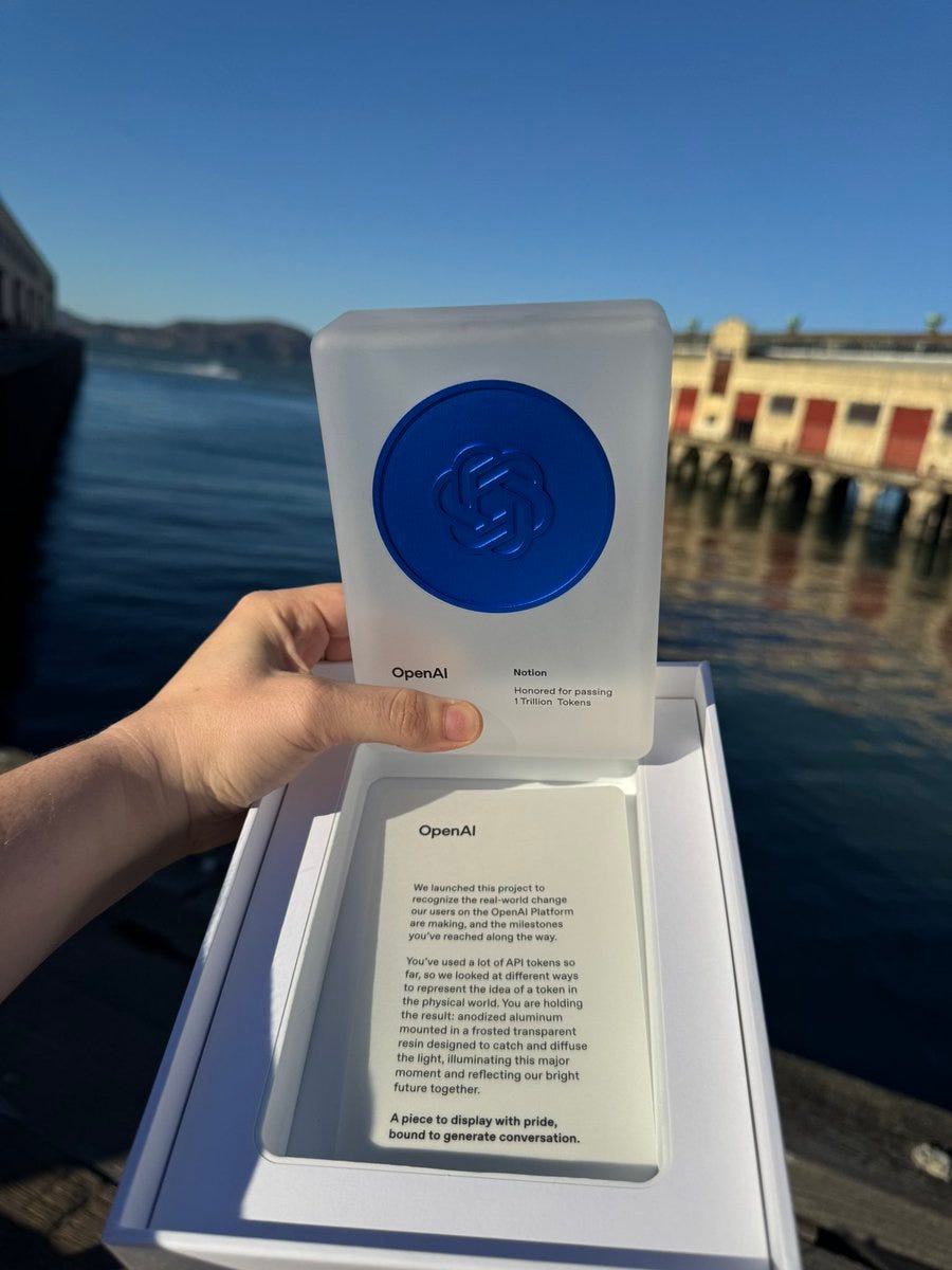 A hand holds a clear acrylic award plaque with a blue circular seal featuring a design, placed on a white base inside an open box with printed text describing the award for processing a trillion tokens on OpenAI, set against an outdoor background of a waterfront with red buildings, blue water, and a clear sky.