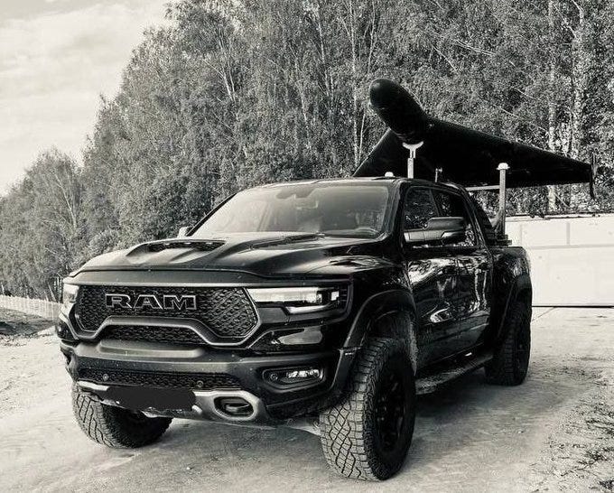 A black RAM truck with a large grille and off-road tires, parked on a dirt path. A black drone, identified as KROOK-1, is mounted on a rack above the truck’s bed. Trees and a building are visible in the background. A black RAM truck with a large grille and off-road tires, parked on a dirt path. A black drone, identified as KROOK-1, is mounted on a rack above the truck’s bed. Trees and a building are visible in the background.