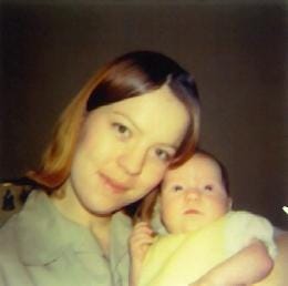 Color photograph from 1970 of Shirley, a young woman with auburn hair, gently holding her infant daughter (the author). Both are looking slightly off camera under warm indoor lighting.