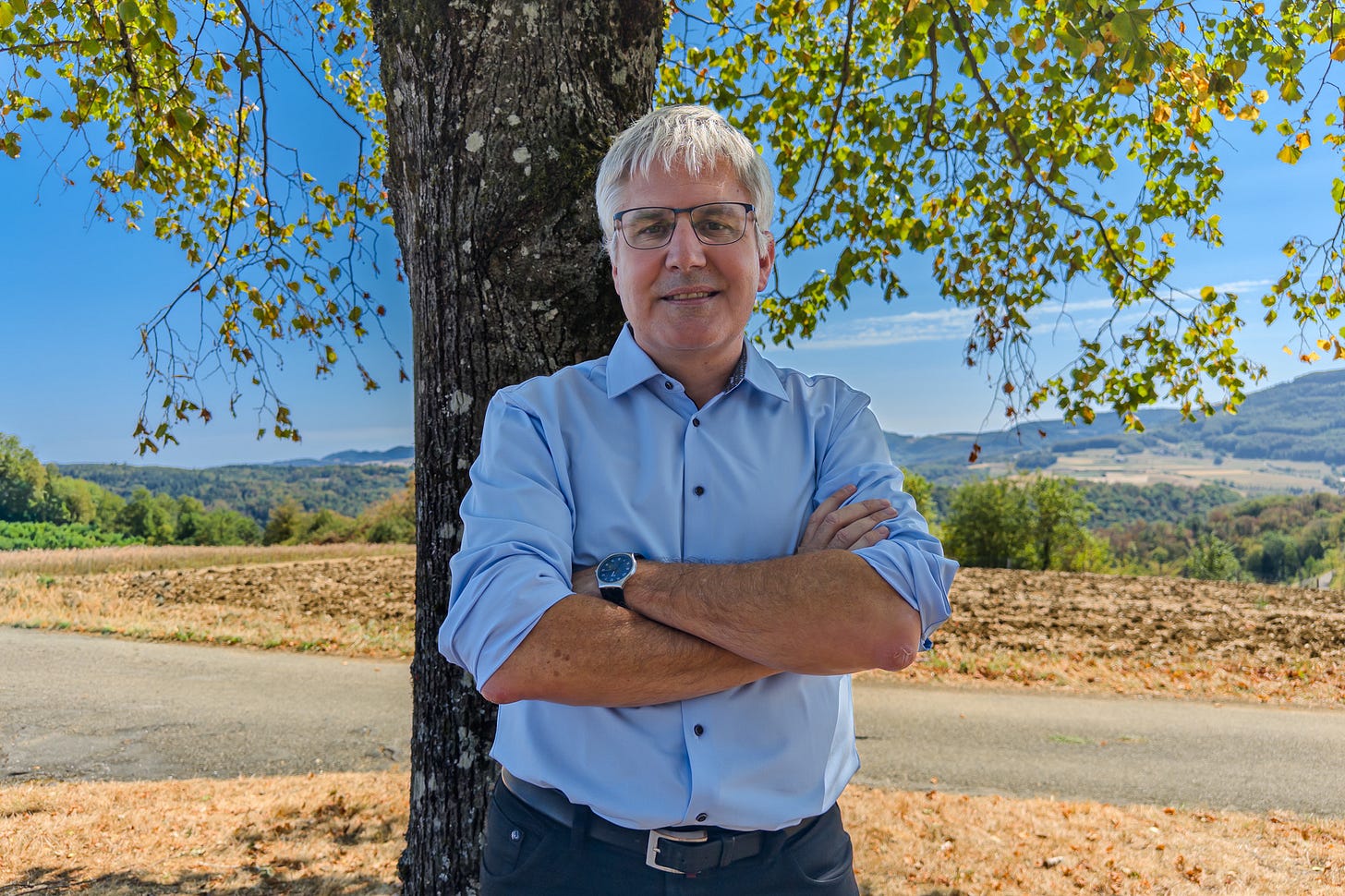 Christian Hofer, Direktor des Bundesamts für Landwirtschaft BLW, steht mit verschränkten Armen unter einem Baum. Hinter ihm liegen ein abgeerntetes Feld und die trockene, hügelige Landschaft unter blauem Himmel.