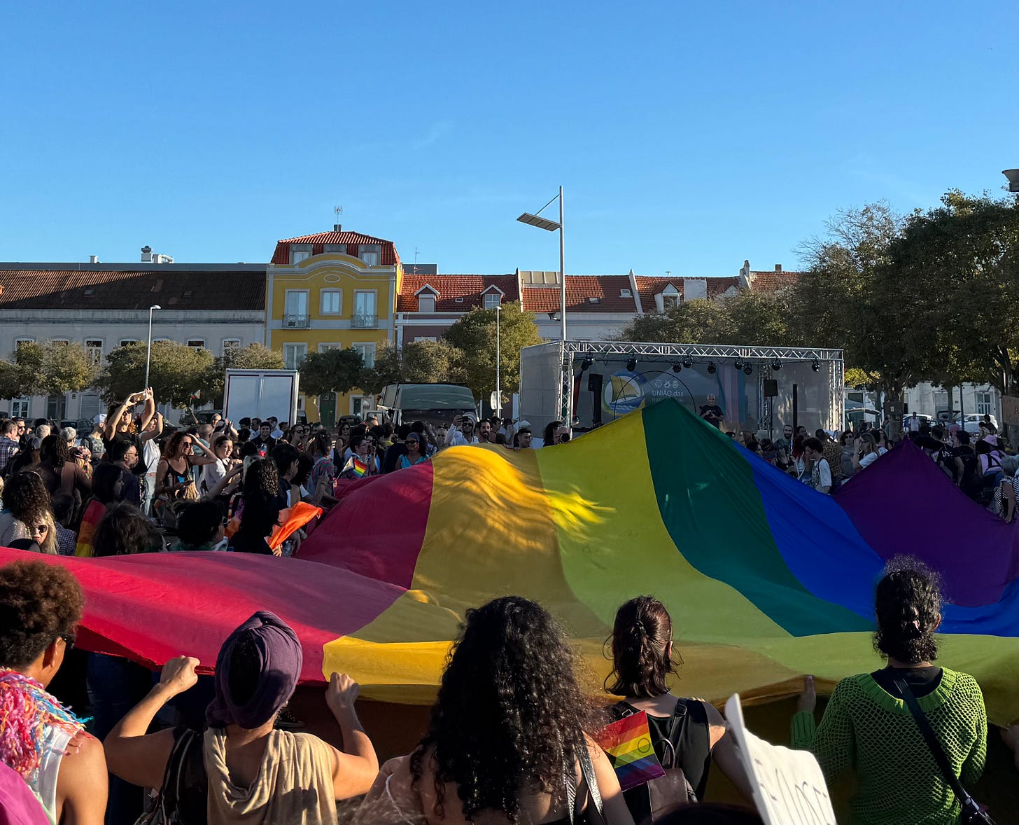 A crowd with a giant rainbow flag in a park.