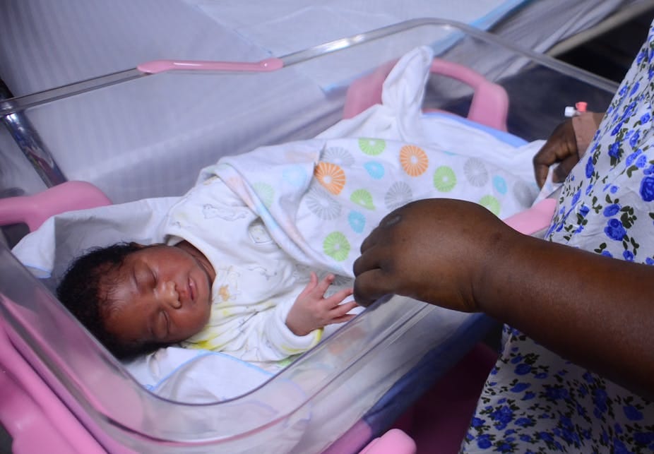 A newborn baby swaddled in a blanket, lying in a bassinet while an adult holds one of its small hands