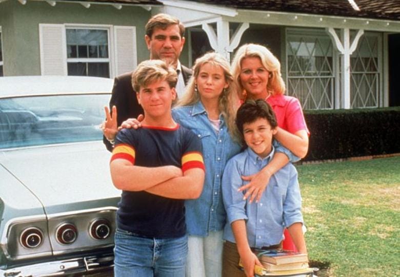 Family portrait of the Arnold family from the sitcom "The Wonder Years." Five people are posed together in the driveway: a man in a dark blazer stands in the back, a woman in a bright pink top stands behind, a blonde boy in front wearing a dark shirt with red and yellow stripes has his arm crossed, a girl in the middle wearing a denim shirt, and a younger boy in a light blue shirt in front right. A car is visible on the left side of the frame, and the house features a green exterior with white trim and latticed windows.