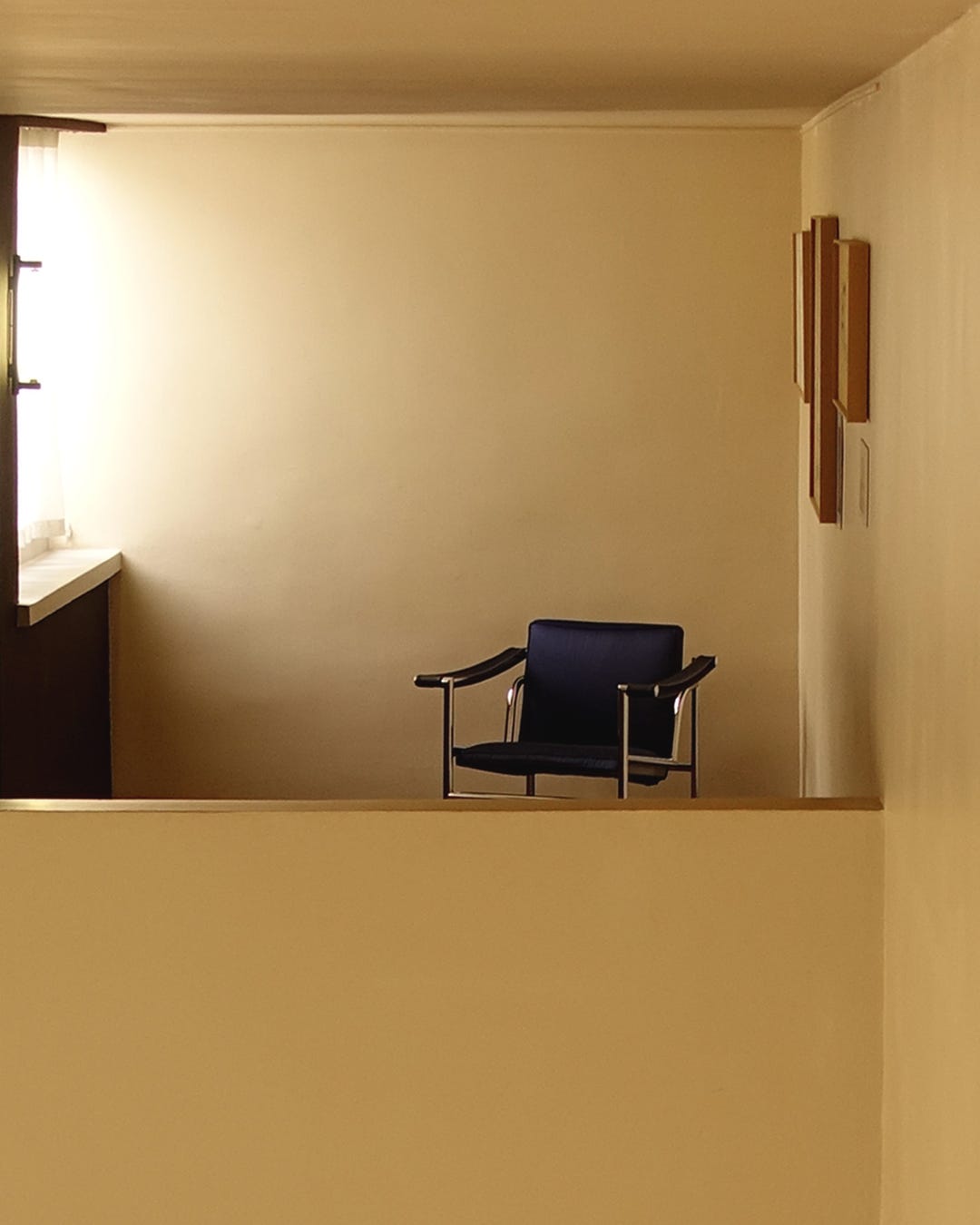 Minimalist interior space with warm cream and tan tones featuring a single black leather chair, wooden wall sculpture, and clean architectural lines creating a serene, contemplative atmosphere