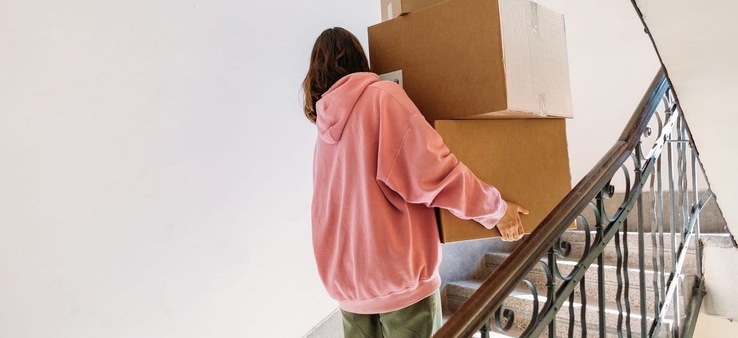 Back view of a woman in a pink hoodie carrying large boxes up a flight of stairs