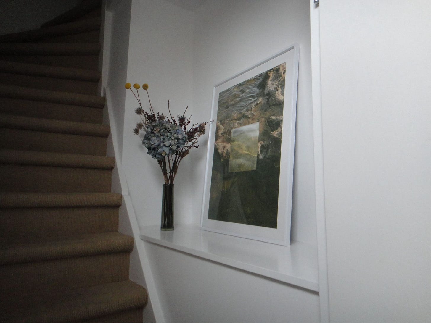 A narrow stairway leading up, with an alcove containing dry flowers and a framed painting.