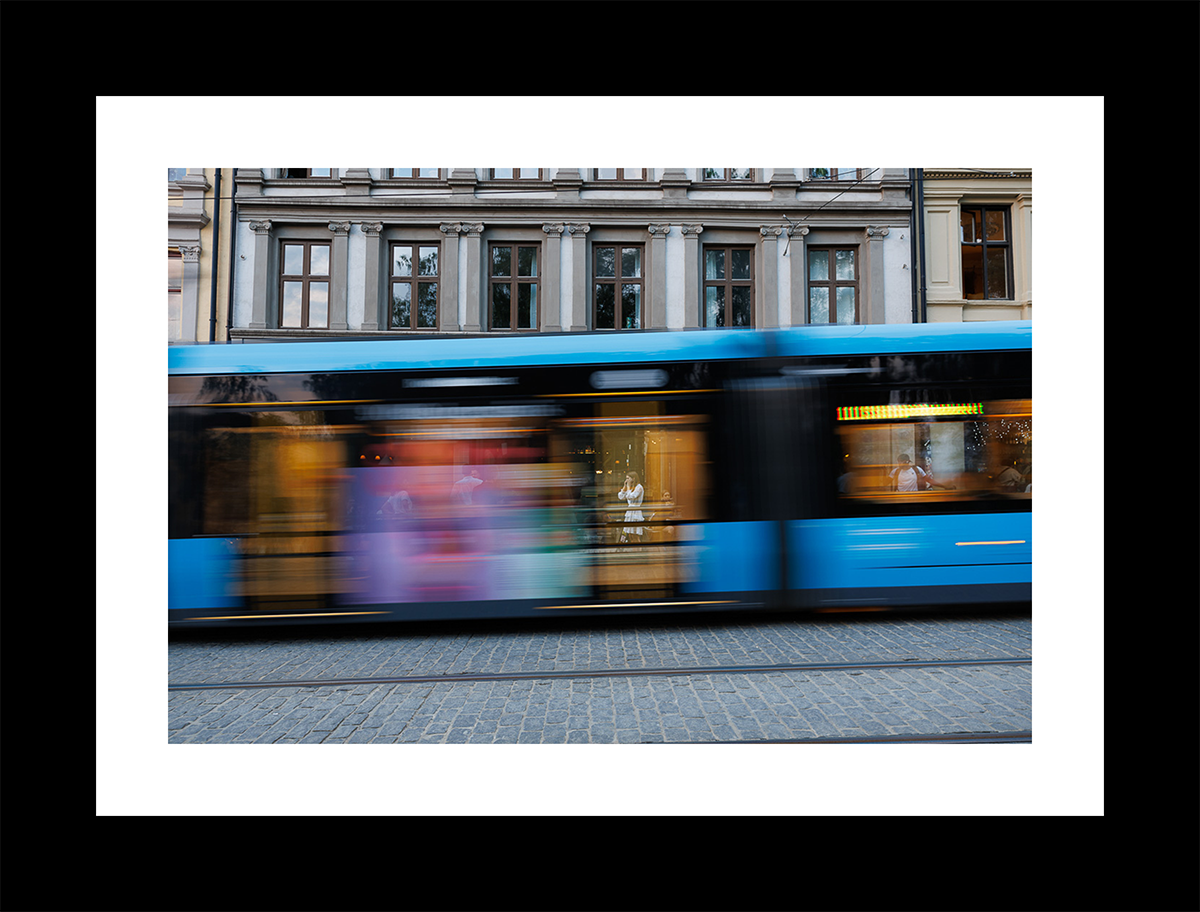 Blue tram in Oslo with motion blur revealing a woman in white seated behind a café window.