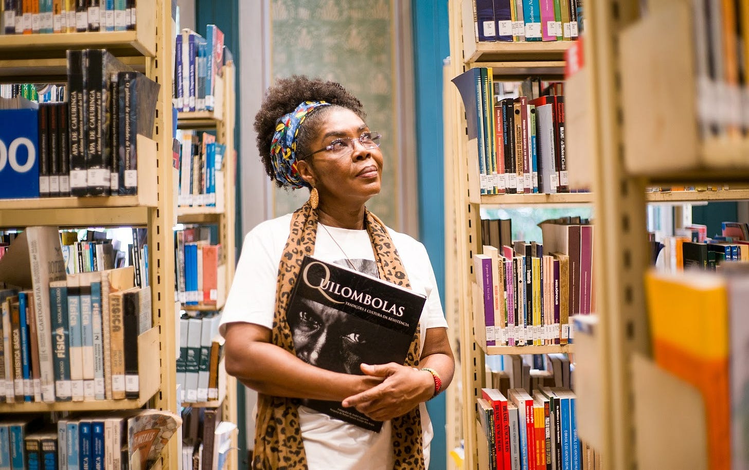 Eli Odara está em pé entre estantes de uma biblioteca. Segura um livro de capa preta, com o título “Quilombolas”, e olha para cima com expressão serena e atenta.