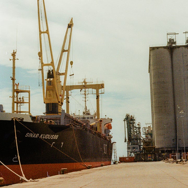 Large cargo ship docked at the Port of Makassar - A massive ship, named "Sinar Kudus88," docked at the industrial Port of Makassar under bright skies, with industrial silos in the background.  Man signaling beside a large cargo ship - A man standing near a motorbike, signaling towards a large docked ship at the Port of Makassar, with the letters "SLT" visible on the ship’s hull.  Truck driver resting in the cab of an orange truck - Close-up shot of an orange truck with a driver’s bare feet visible, resting in the cabin at the Port of Makassar.  Truck with open door and driver relaxing - An orange truck with its door wide open, the driver’s legs stretched out of the cabin in a relaxed posture while parked at the port.  Truck parked at the port near industrial silos - A truck labeled "Karawa" parked at the dockside in the Port of Makassar, industrial buildings and silos in the distance behind.