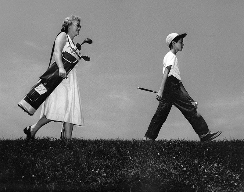 boy walking with golf club with mom carrying his bag, walking behind boy walking with golf club with mom carrying his bag, walking behind