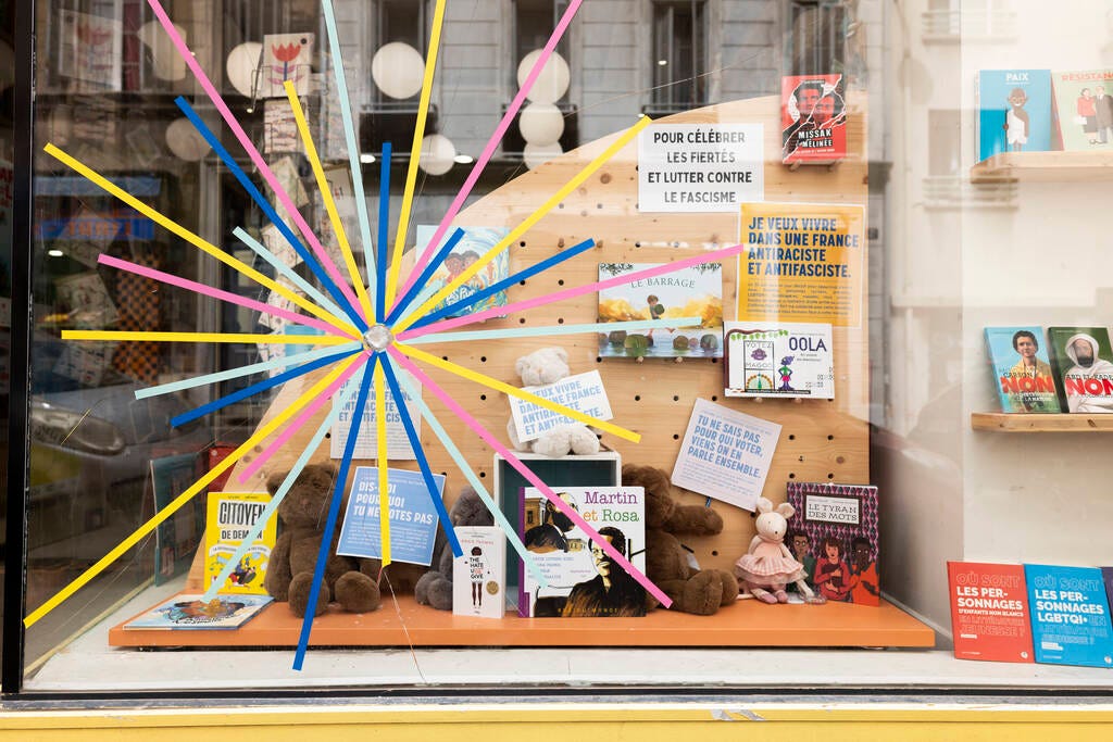 Vitrine de la librairie Pantagruel à Marseille. On voit un impact, et des bouts de scotch colorés, placés comme des rayons autour de l'impact. Derrière la vitre, des peluches qui tiennent des pancartes et divers livres engagés. 