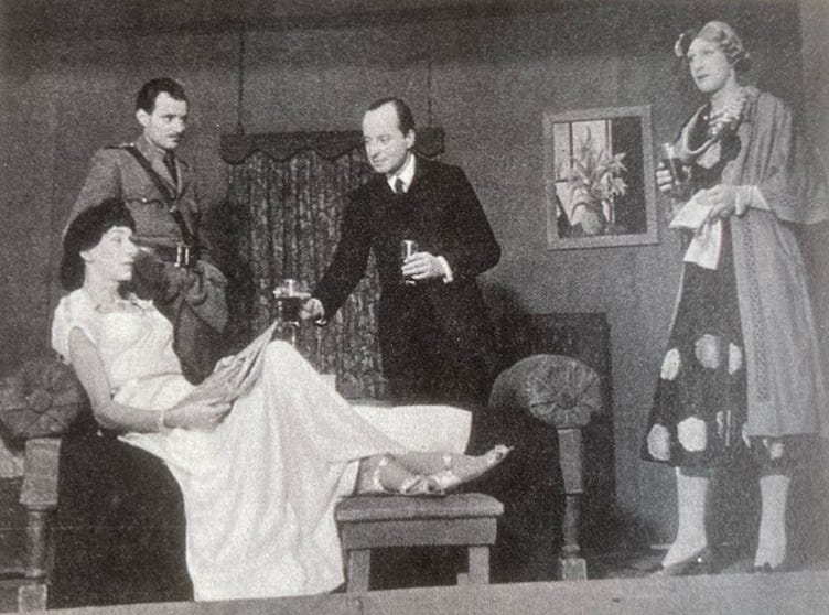 A 1920s cocktail party, staged in a WWII prison camp. A man in a suit hands out drinks, while another man in uniform stands by a chaise longue on which a man wearing a wig and white gown reclines with a magazine. Standing up further across the stage is another man in a dress, wig and hat